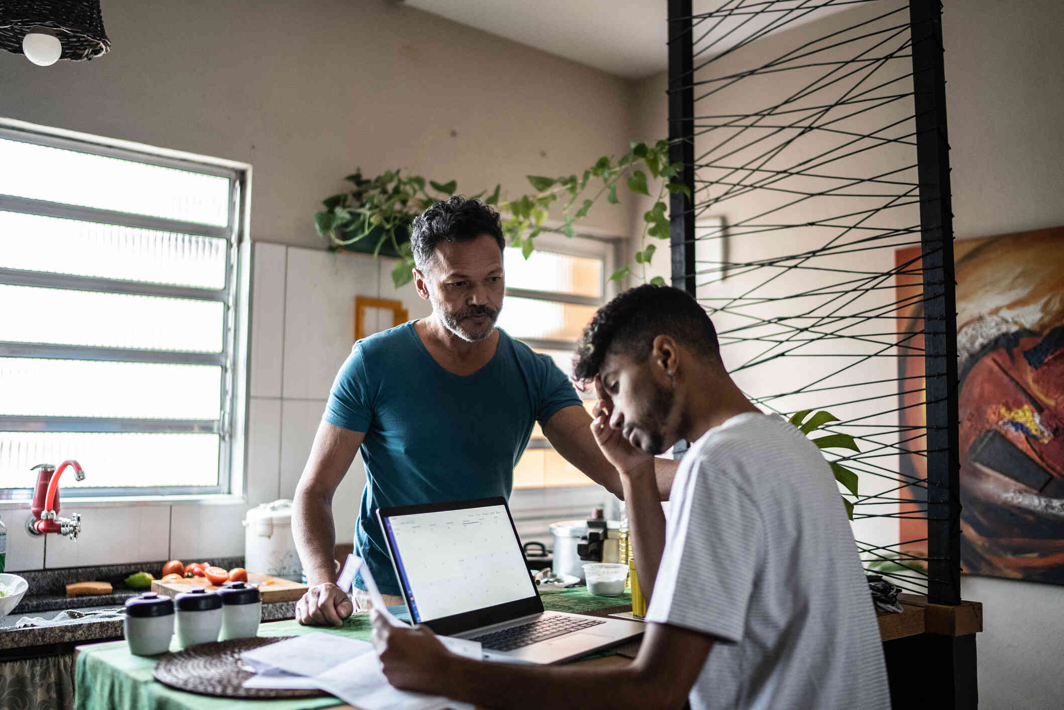 A man sits at a kitchen counter with his laptop open infront of him with a troubled expression as his male partner stands infront of him looking sad.